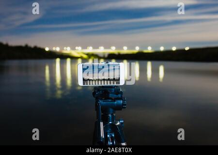 Mit Smartphone wie professionelle Kamera auf Stativ und dem Erfassen von Nacht Landschaft mit viel Licht auf der Oberfläche des Wassers wider Stockfoto