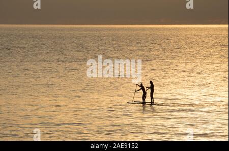 Eine Frau und ein mit Stand up Paddleboards außerhalb Lyme Regis Hafen Anfang Dezember bei Sonnenaufgang Mann. Lyme Regis ist auf der Heritage Coast gelegen ist oder Stockfoto