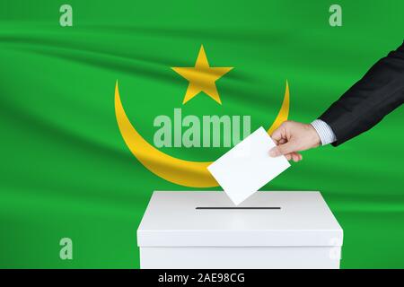 Wahlen in Mauretanien. Die Hand des Menschen, der seine Stimme in die Wahlurne setzt. Mauretanien Flagge auf Hintergrund gewellt. Stockfoto