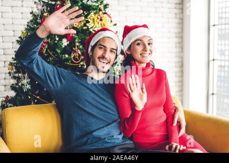 Portrait von Happy süßes Paar in santa Hüte Spaß an der Kamera Lächeln, winken und Sagen hallo Sie und genießen Sie die Zeit zusammen, in Christus zu Stockfoto