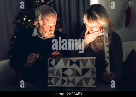 Älterer Mann, Geschenk, um Frau und Spaß Eröffnung Magic Christmas box sitzen zusammen auf dem Sofa im Wohnzimmer zu Hause. Ruhestand Paar anhand von quantitativen Simulatio Stockfoto