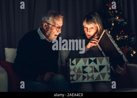 Älterer Mann, Geschenk, um Frau und Spaß Eröffnung Magic Christmas box sitzen zusammen auf dem Sofa im Wohnzimmer zu Hause. Ruhestand Paar anhand von quantitativen Simulatio Stockfoto