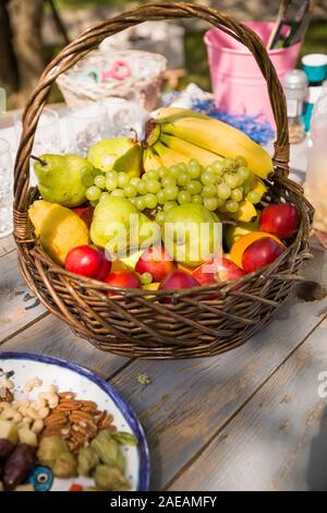 Korb mit Obst (Äpfel, Birnen, Bananen und Trauben) auf dem Holztisch mit Muttern und andere ingridients für putdoor Picknick. Gesunde Lebensmittel für Stockfoto