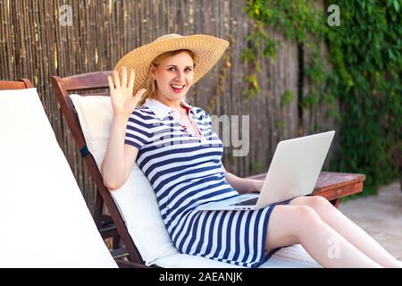 Hallo! Portrait von positiven junge erwachsene Freiberufler Frau in Hut und Kleid sitzt auf gemütliche Liege mit Laptop und mit fünf Geste mit eine Stockfoto