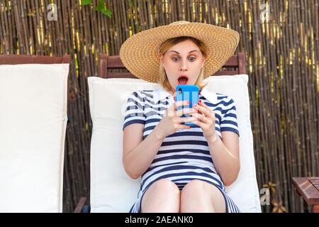 Portrait von überrascht junger Erwachsener blogger Frau in Hut und Kleid sitzt auf Sonnenbank und Holding Telefon, Bildschirm News lesen Mit schockiert Fa Stockfoto