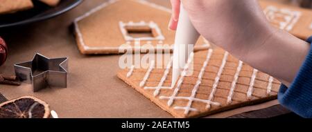 Frau verziert Lebkuchen cookies Haus mit weißer Glasur Puderzucker sahne Topping auf Holztisch, Hintergrund, Backpapier in Küche, Nahaufnahme, m Stockfoto