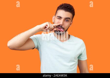 Portrait von Silly kindisch brünett Mann mit Bart in lockeren weißen T-Shirt seine Nase bohren und Zunge mit törichten komische Gesicht, schlechte Manieren. Stockfoto