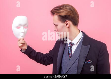 Portrait von umgekippt schwerer junge Unternehmer im eleganten Anzug und mit stilvollen Haarschnitt aufmerksam auf weiße Maske mit düsteren gedrückt expressio Stockfoto
