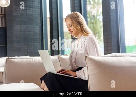 Portrait von attraktiven Heiter erfolgreiche Frau mit blonden Haaren in der klassischen Kleidung sitzen auf bequemen Couch und Arbeiten am Laptop, Sie tun b Stockfoto