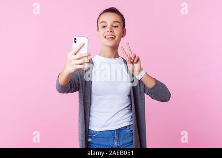 Portrait von fröhlichen Verrückten jugendlich Mädchen mit bun Frisur in Freizeitkleidung Gestik Sieg unterzeichnen und mit Zunge, während das Video Anruf am Telefon. Stockfoto