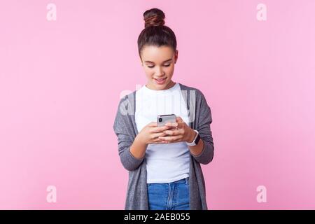 Portrait von Happy schönes junges Mädchen mit bun Frisur in Freizeitkleidung mit Handy, Gefühl aufgeregt Plaudern mit Freunden, so scrollen Stockfoto