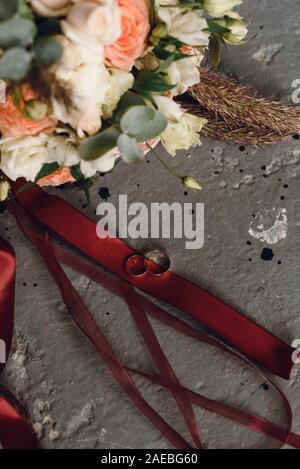 Ringe der Jungvermählten auf einem grauen grunge Hintergrund und der Hintergrund einen Blumenstrauß der Bräute. Stockfoto