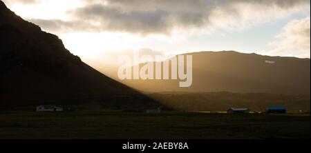 Schönen Abend Licht mit der Sonne zwischen den Wolken und Berge glänzen, das Dorf von Arnarstapi im Vordergrund, Island. Stockfoto