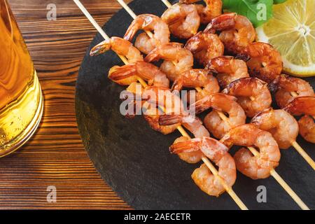 Garnelen gebraten am Spieß mit Rucola, Zitrone auf einem schwarzem Schiefer Teller und Bier in einem Glas auf einen hölzernen Tisch. Stockfoto