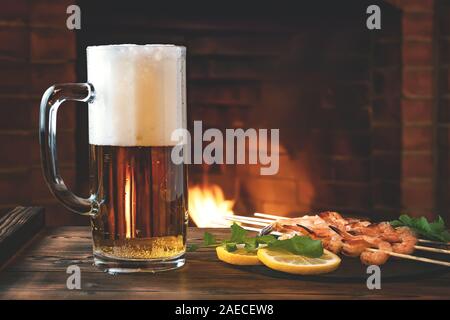 Becher Bier, gebraten auf Spieße mit Garnelen mit Rucola Zitrone auf einem schwarzem Schiefer Teller auf einen hölzernen Tisch in ein Zimmer mit Kamin. Stockfoto