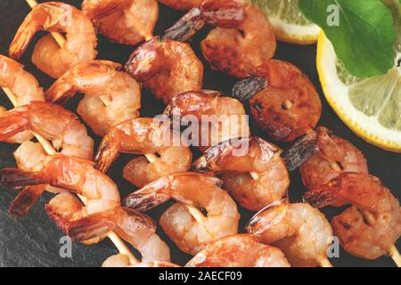 Garnelen gebraten am Spieß mit Rucola und Zitrone auf einem schwarzem Schiefer Teller auf einem Holztisch, Ansicht von oben, flach, aus der Nähe. Stockfoto