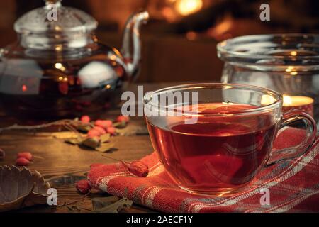 Tee mit Weißdorn in einem Glas Schale und Teekanne mit einem Teelicht auf einem Holztisch, in einem Raum mit einer brennenden Kamin. Stockfoto