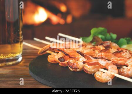 Gebraten auf Spieße mit Garnelen mit Rucola Zitrone auf einem schwarzem Schiefer Teller, Becher Bier auf einer hölzernen Tisch in Zimmer mit Kamin. Stockfoto