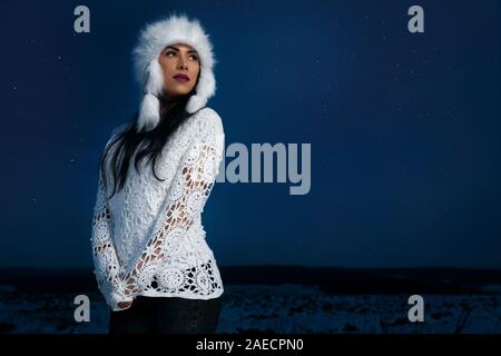 Modische Modell tragen im stilvollen warme Mütze und weißem gestrickt Bluse posiert auf dem Feld auf dem Hintergrund der Nachthimmel im Winter. Schöne Mädchen mit schwarzen langen Haaren stehen, entfernt. Stockfoto