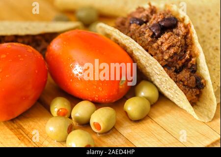 Frische taco Zutaten rote reife Pflaumen Tomaten, grüne Oliven und Rind Tacos mit schwarzen Bohnen in Taco shells auf einer hölzernen Schneidebrett. Stockfoto