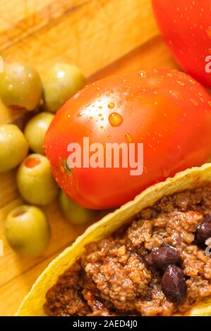 Frische taco Zutaten rote reife Pflaumen Tomaten, grüne Oliven und Rind Tacos mit schwarzen Bohnen in Taco shells auf einer hölzernen Schneidebrett. Stockfoto