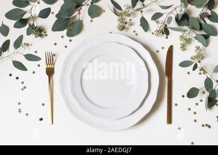 Weihnachten, Neujahr Tisch setzen. Golden Besteck, Porzellan Teller, Eukalyptus, Zweige und golden Konfetti Sterne auf weißem Hintergrund Stockfoto