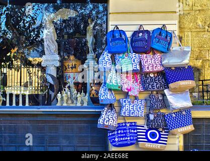 Monastiraki, Athen/Griechenland - 06 Dezember 2019: Taschen mit Griechenland auf ihren Seiten außerhalb der Souvenirshop hängenden gedruckt. Statuen Replikate und Reflexionseigenschaften Stockfoto