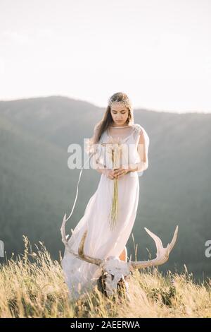 Schöne Mädchen Braut in einem langen Kleid, Wicklung im Wind Haarschmuck im Profil, Draußen, Berg, deer Skull mit geweih an den Füßen weddi Stockfoto