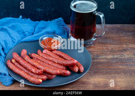 Geräucherte Würstchen auf einem dunklen Platte. In der Nähe von einem großen Glas dunkles Bier. Kopieren Sie Platz. Stockfoto