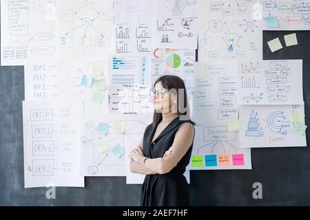 Junge erfolgreiche weibliche Ökonom mit verschränkten Armen durch die Tafel stehend Stockfoto