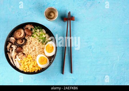 Ramen. Soba Nudeln mit Ei, Shiitake und enoki Pilze, Mais und Frühlingszwiebeln, Overhead shot auf blauem Hintergrund mit Stäbchen, Sake Stockfoto