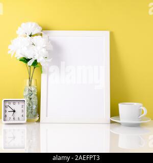 Weißer Rahmen, Blumen in Vase, Schale mit Tee oder Kaffee, Uhr auf einem weißen Tisch gegen gelbe Wand mit kopieren. Mock up. Stockfoto