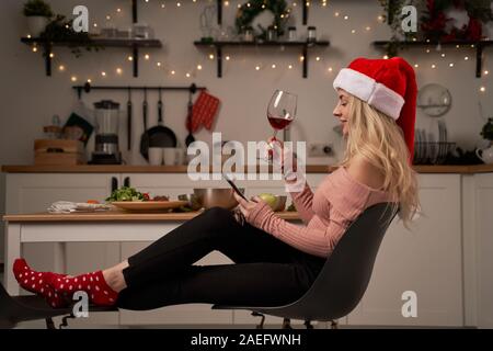 Mädchen in santa hut mit Weinglas und Telefon sitzt in der Küche mit brennenden Girlanden. Das Konzept des Neuen Jahres Stockfoto