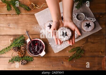 Sternförmige Weihnachtsplätzchen auf einen hölzernen Tisch. Biquites mit Marmelade und Pulver, Dekor aus Zweigen Fichte. Kinder Hand in den Rahmen. Blick von oben. Platz kopieren Stockfoto