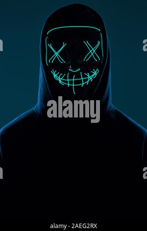 Porträt eines anonymen Mann in einem schwarzen Kapuze sein Gesicht versteckt sich hinter einem furchtsamen neon Mask. Studio gedreht. Stockfoto