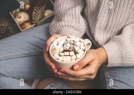 Eine Frau in einem gestrickten Pullover in ihren Händen hält Kakao mit Marshmallows und Schokolade in einem gemütlichen Zuhause. Stockfoto