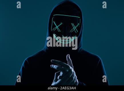 Porträt eines anonymen Mann in einem schwarzen Kapuze sein Gesicht versteckt sich hinter einem furchtsamen neon Mask. Studio gedreht. Stockfoto