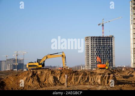 Bagger auf einer Baustelle auf dem Hintergrund einer Baukräne und Gebäude. Tieflöffel graben den Boden für die Stiftung, Festlegung Sturmabwasserkanal Stockfoto