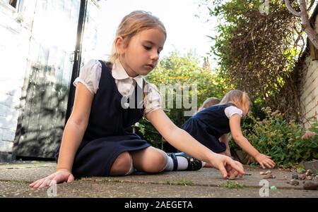 Ein kleines Mädchen in Schuluniform sitzt auf dem Boden Tief in Gedanken, die mit ihrer Schwester im Garten spielen Im Hintergrund Stockfoto