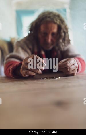 Senior obdachlosen Mann mit lockigem Haar und zerrissenen Pullover Zählen von Münzen auf hölzernen Schreibtisch in einem konzeptionellen Bild der Armut und Krisen. Stockfoto