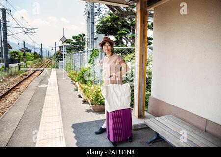 Japanische Frau mit Hut stehend auf Bahnhof Plattform mit Einkaufstasche und rosa Koffer. Stockfoto