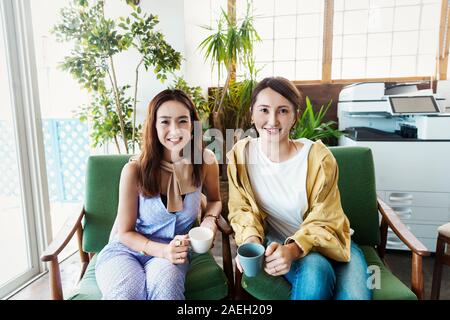 Zwei weibliche Japanische Fachleute sitzen in einem Co-Arbeitsraum, ein Lächeln auf die Kamera. Stockfoto