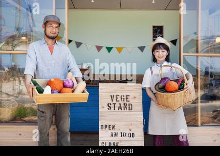 Japanischer Mann und Frau, die außerhalb einer Farm Shop, Kiste und Korb mit frischem Gemüse, an der Kamera schaut. Stockfoto