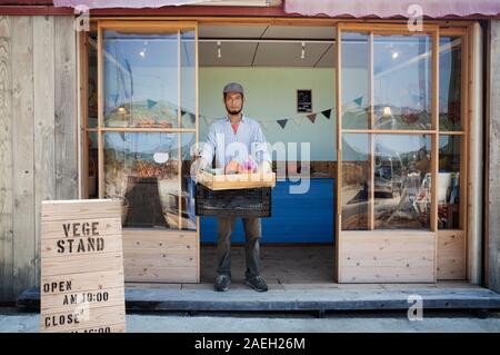 Japanischer Mann außerhalb einer Farm Shop, die hielten Kiste mit frischem Gemüse, an der Kamera schaut. Stockfoto