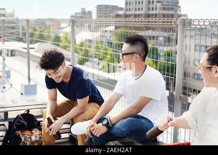Drei junge japanische Männer sitzen auf einer Dachterrasse in einem urbanen Umfeld, trinken Bier. Stockfoto