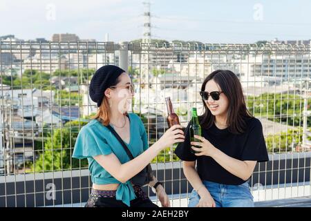 Zwei junge japanische Frauen sitzen auf einer Dachterrasse in einem urbanen Umfeld, trinken Bier. Stockfoto