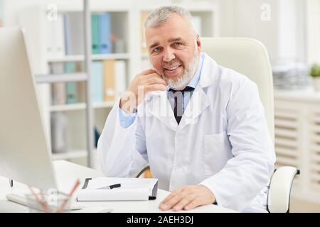 Portrait von Reifen Doktor mit grauem Haar und Bart lächelnd an Kamera, während an seinem Arbeitsplatz im Büro Stockfoto