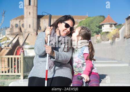 Blinde Mutter mit einem weißen Stock und ihre Tochter sprechen, sitzen auf einer Bank im Park und lächelnd. Sehbehinderte lifestyle Konzept. Stockfoto