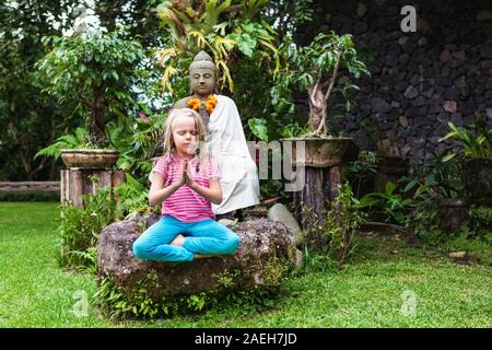 Morgen lustig Meditation in den tropischen Garten. Glückliches Mädchen sitzen, im Yoga Lotus entspannen Pose mit Mudra namaste Hände. Aktives Kind gesunder Lebensstil, Reisen Stockfoto