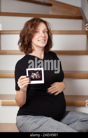 Schwangere curly Frau ihr Baby Sonographie. Glücklich werdende Dame genießen erste Foto von ihrem ungeborenen Kind, ihr zukünftiges Leben zu antizipieren, kopieren Stockfoto